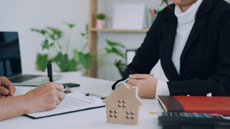 A person reviews a homeowners insurance document with an agent at a desk with a house model and paperwork