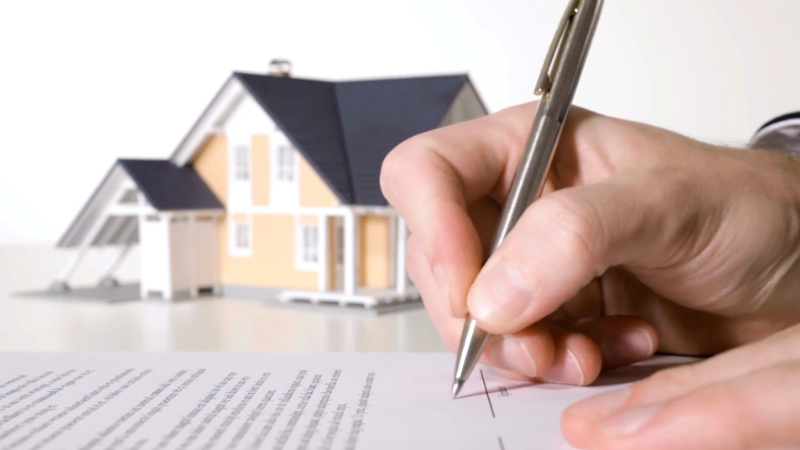 A person signs a home insurance paper with a house model nearby