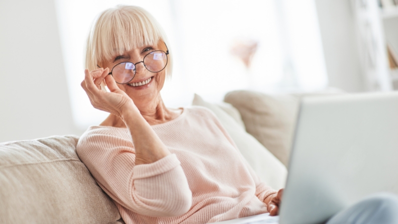 A smiling older woman sits on a sofa using a laptop to learn about average retirement ages worldwide