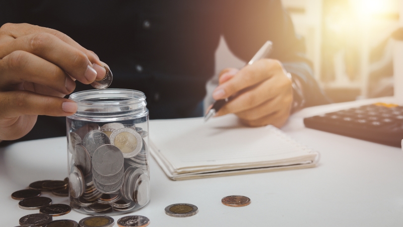 A person adds coins to a jar while taking notes and using a calculator to track savings for retirement expenses