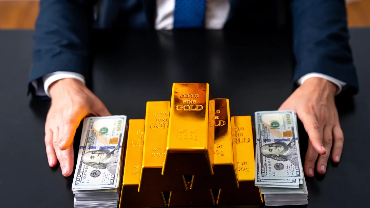 A businessperson sits behind gold bars and stacks of U.S. dollar bills