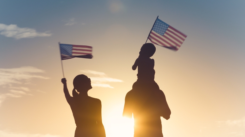 A family stands together at sunset holding American flags, with a child sitting on an adult’s shoulders