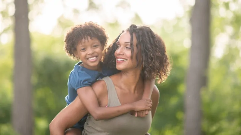 A mother carries her young child on her back outdoors, reflecting family bonds in a time of fewer babies and changing demographics