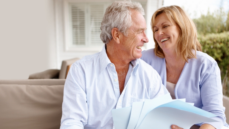 A smiling older couple sits together holding paperwork as they plan for retirement