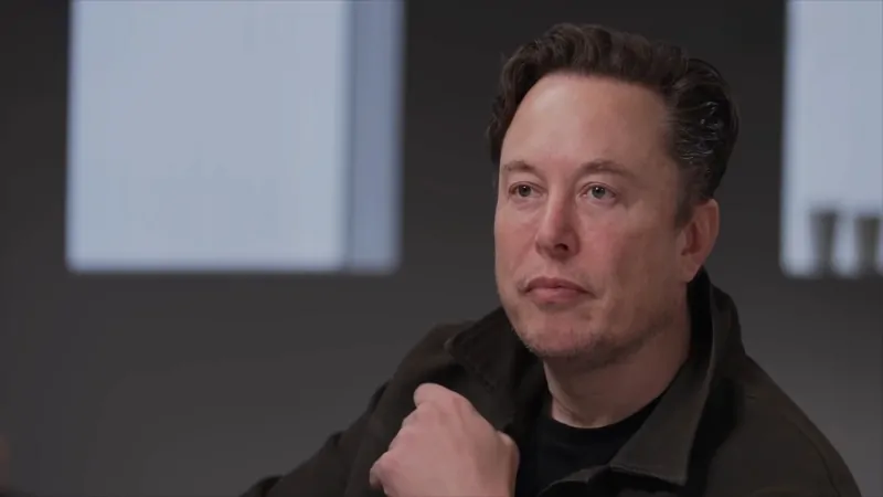 Elon Musk sits thoughtfully in a dark jacket during an interview