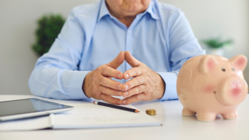 An older man sits at a desk with a piggy bank, tablet, and notes as he reviews his retirement planning by age group