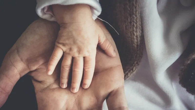 An adult hand holds a baby’s hand, symbolizing support and concern about high child care costs