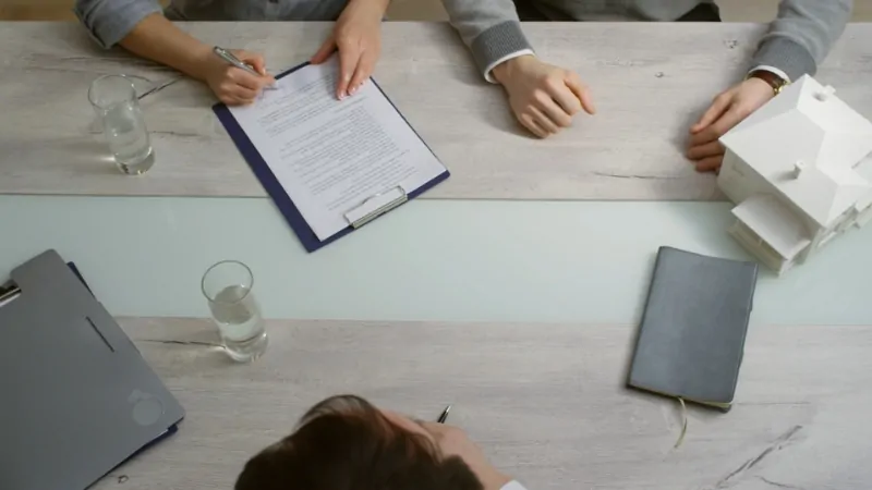 Three people sit at a table discussing housing documents, with a model house and paperwork in view