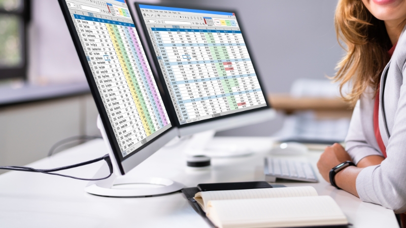 A professional reviews health survey data on two computer monitors filled with spreadsheets and statistics