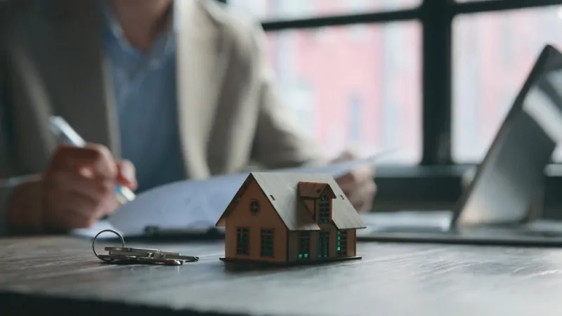 A small model house sits on a desk as someone signs papers in the background, highlighting the link between falling birth rates and housing demand