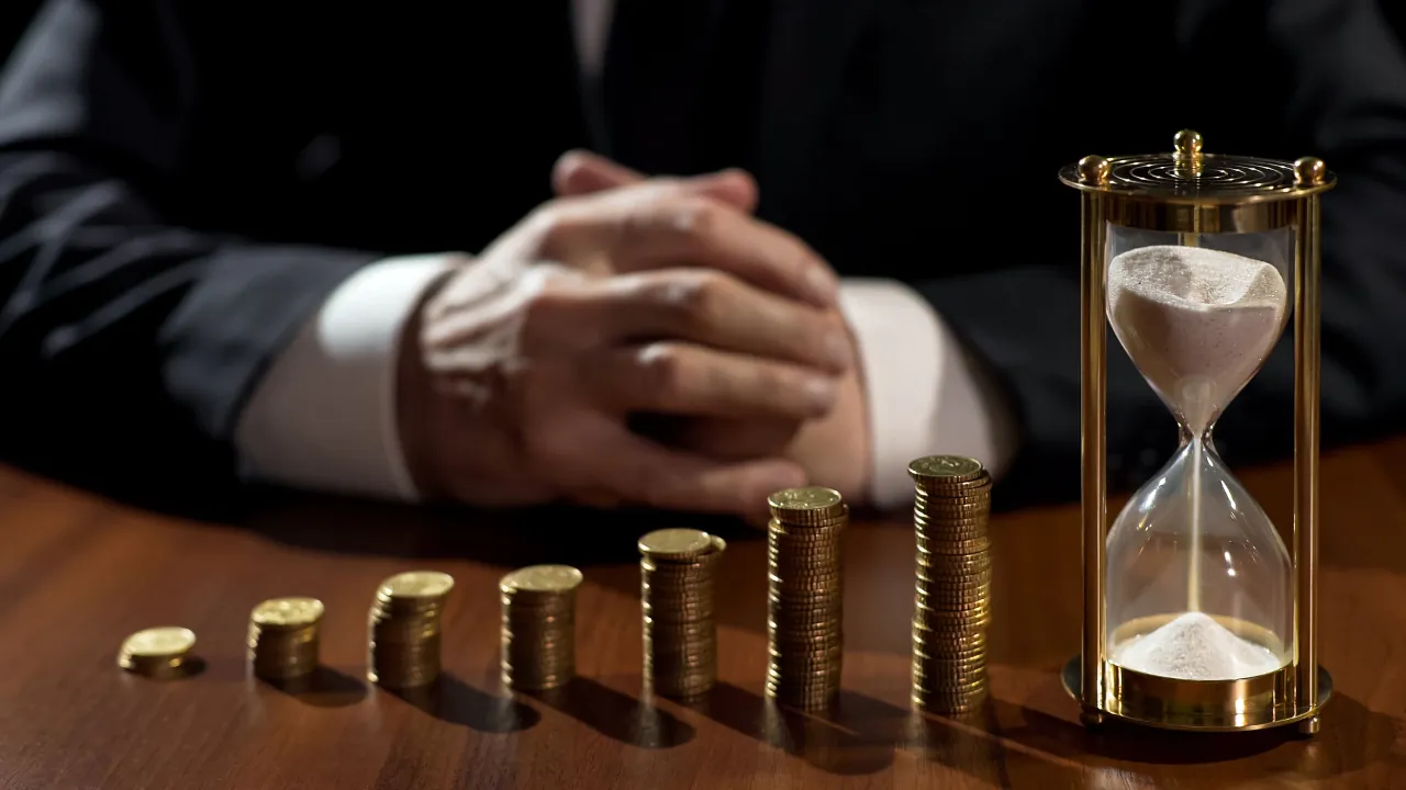 A person in a suit sits behind coin stacks and an hourglass on a desk
