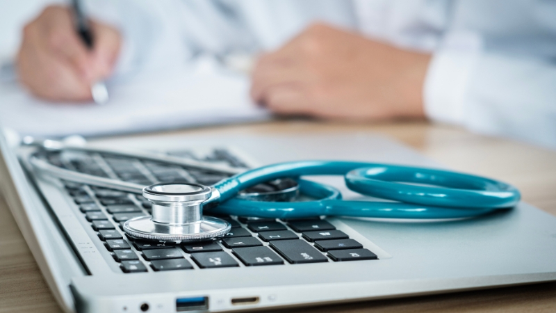 A stethoscope sits on a laptop keyboard while a healthcare professional writes notes in the background