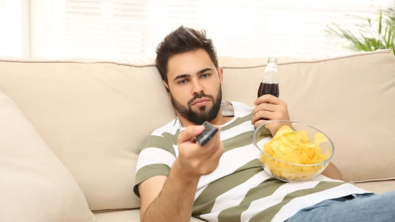 Man on a couch holding a remote, soda, and chips, showing a common example of adult inactivity outside of work
