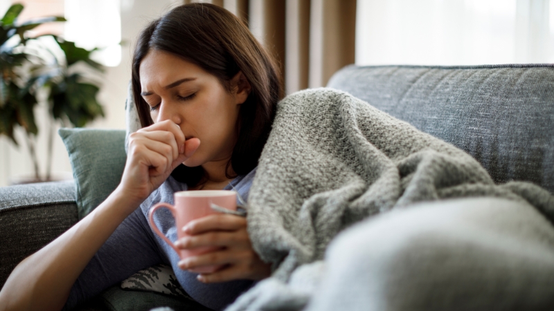 Woman wrapped in a blanket on a couch holding a mug and coughing, showing signs of stomach upset or illness