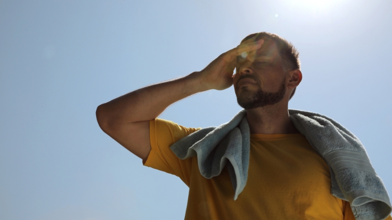 Man in a yellow shirt shielding his eyes from the sun with a towel over his shoulder, showing heat and dehydration risk