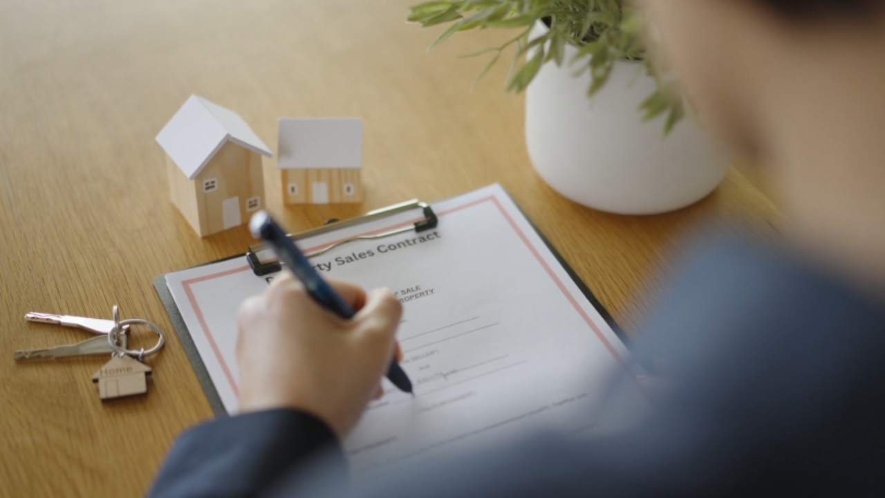 A person signs a property sales contract beside house keys and small home models