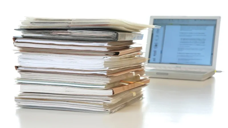 A stack of documents next to a laptop displaying text, representing research for online health articles
