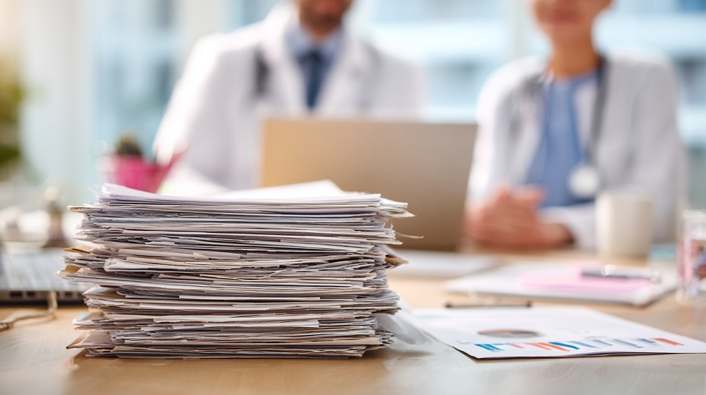 Stack of medical files and documents on a desk in a bright office, with blurred doctors in the background discussing certificate of live birth data