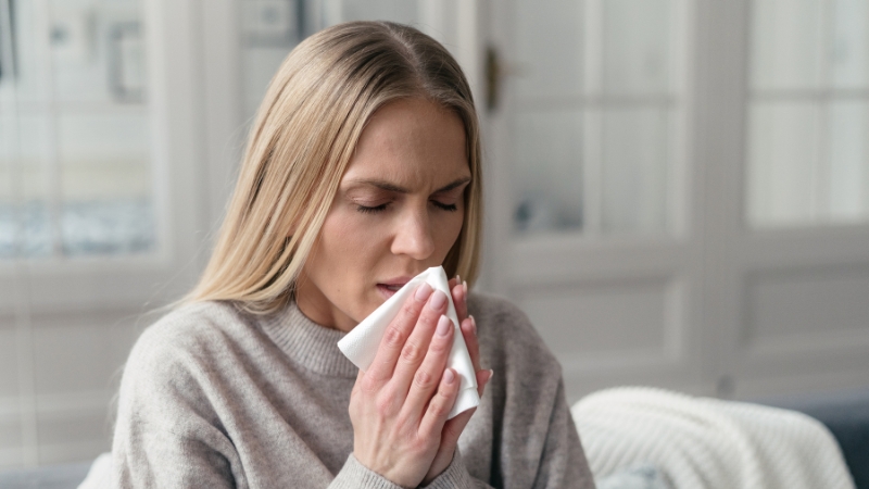 Woman holding a tissue and coughing indoors, showing signs of a seasonal respiratory infection