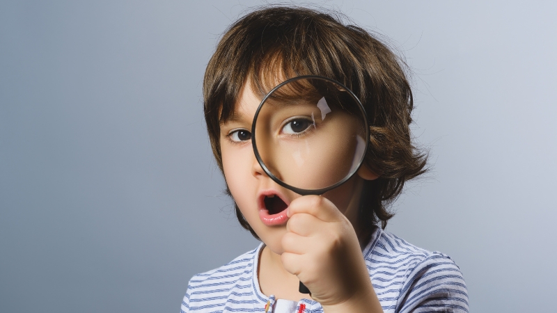 Young child holding a magnifying glass to their eye, looking curious and surprised
