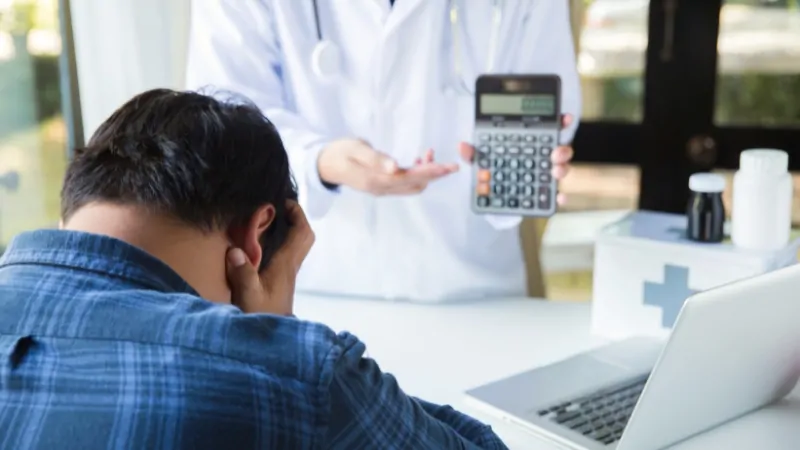 A patient looks stressed as a doctor shows a calculator, reflecting concerns over costs and challenges to the Fair Pricing Act