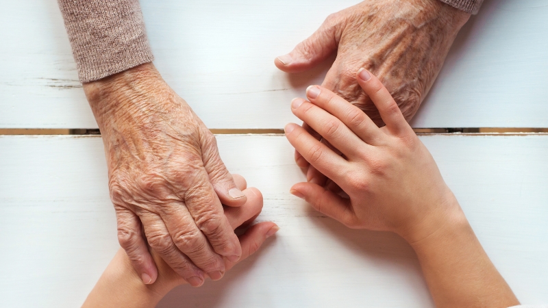 An elderly person’s hands rest on top of younger hands across a table