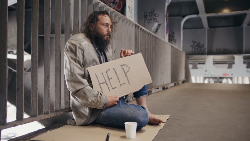 A homeless man sits on cardboard under a bridge holding a sign that says “HELP” with a cup placed in front of him