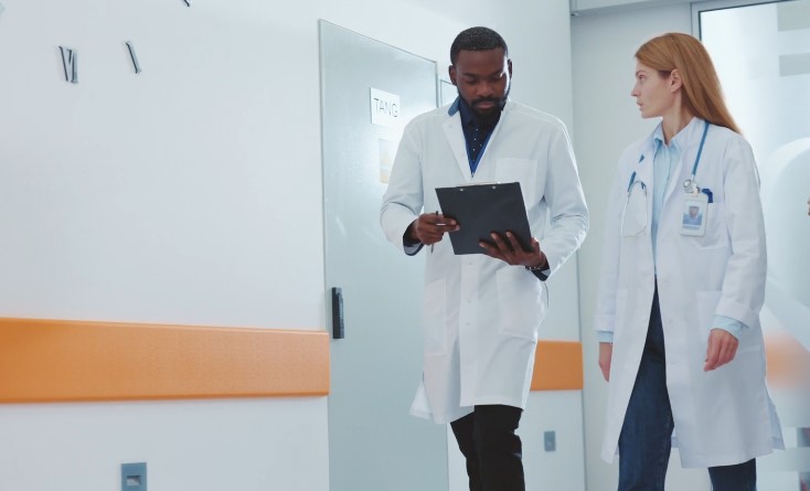 Two medical professionals, a man with a clipboard and a woman with a stethoscope, walk down a brightly lit, sterile hospital hallway
