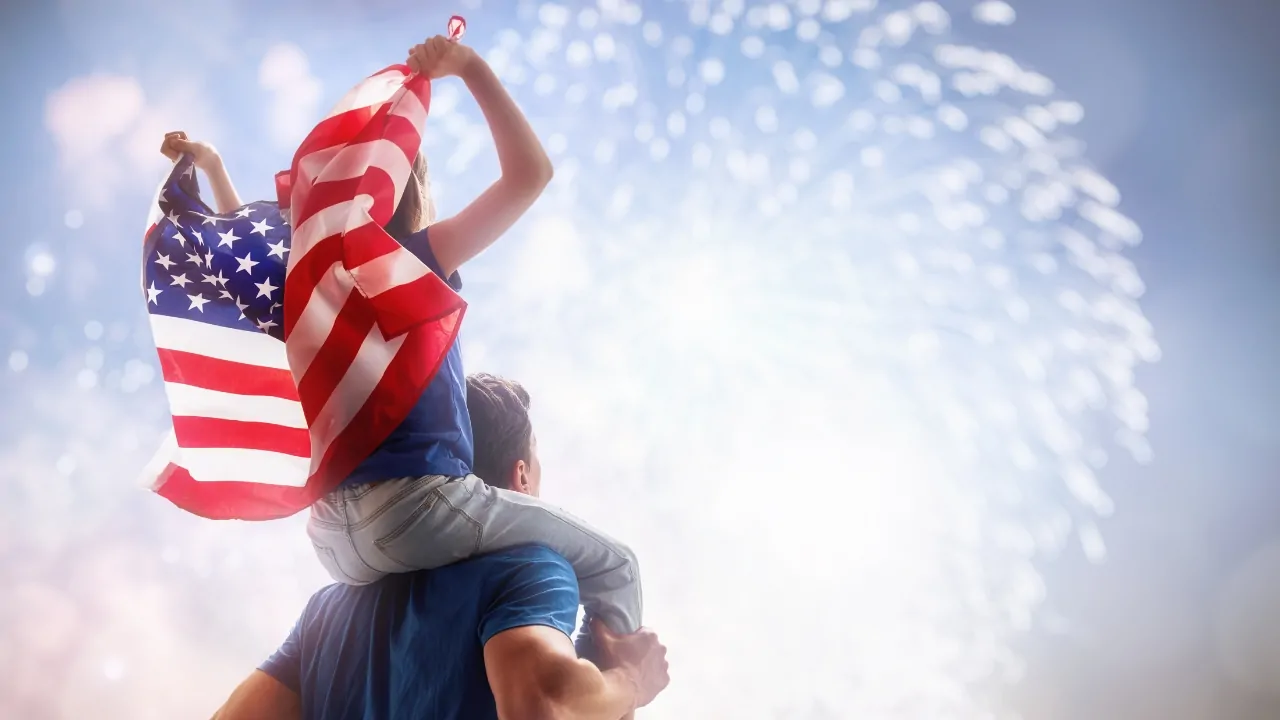 Child waves an American flag on someone’s shoulders during a fireworks celebration