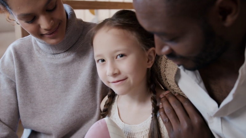 A smiling young girl sits between two adults at home, representing adopted children in the United States