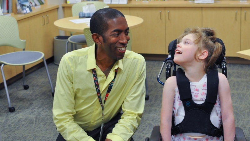 A therapist smiles while interacting with a young girl in a wheelchair during a cerebral palsy therapy session