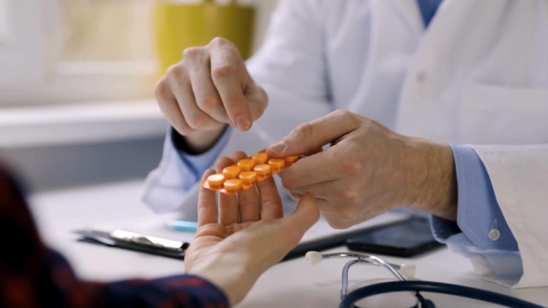 A doctor hands a patient a pack of orange tablets during a checkup