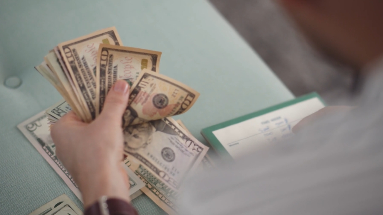 A person counts U.S. cash beside a checkbook on a table