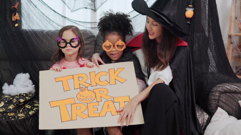 Two kids in Halloween costumes sit with a woman dressed as a witch, holding a handmade “Trick or Treat” sign