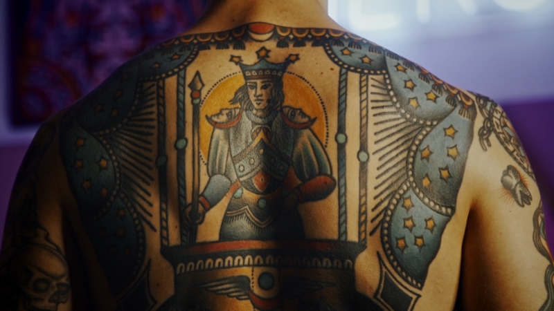 A man with a large back tattoo stands in soft light as lifestyle factors behind cancer risk are discussed