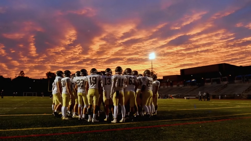 A football team huddles on the field under a vivid sunset sky, preparing for the big game on Super Bowl Sunday