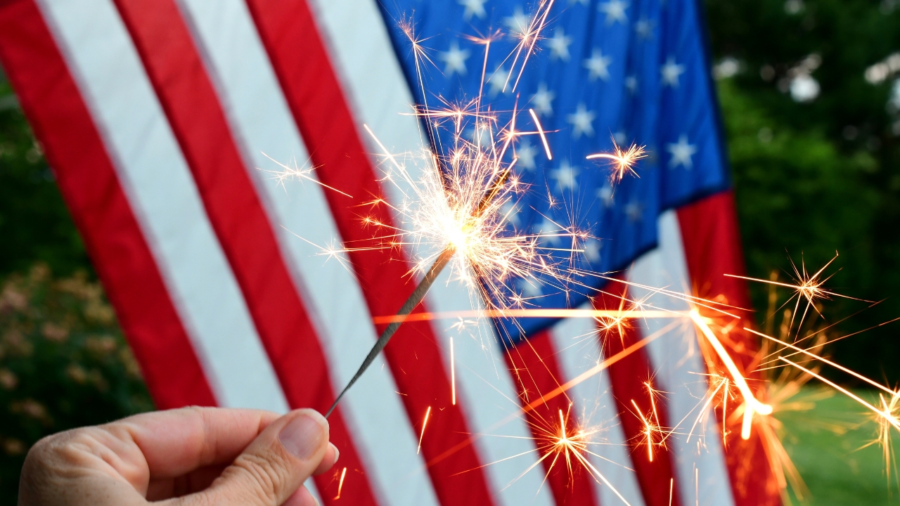 Hand holds a lit sparkler in front of an American flag during a holiday celebration