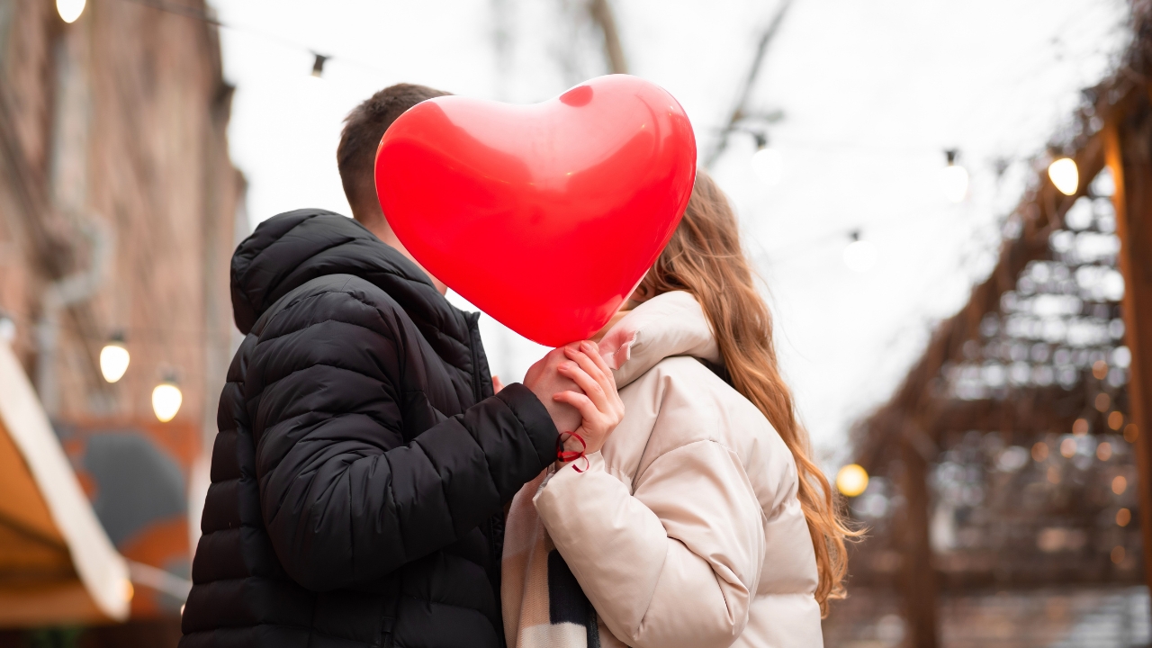 Couple share a Valentine’s Day moment behind a red heart balloon on a city street