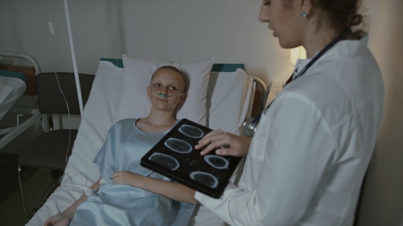 A cancer patient lies in a hospital bed while a doctor shows scan images on a tablet