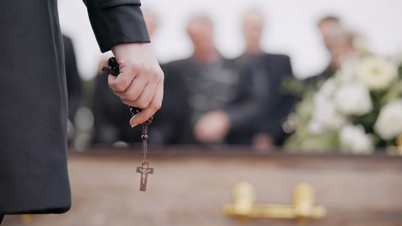 A hand holds a cross at a funeral as mourners stand quietly in the background