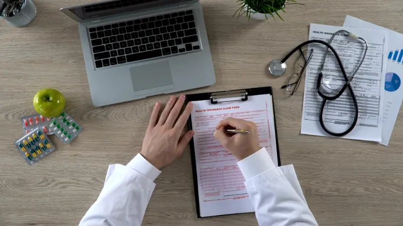 Person fill out a health insurance form at a desk with a laptop, documents, and medical tools