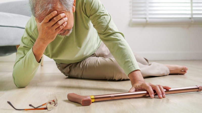 An older man lies on the floor after a fall as he reaches for his cane