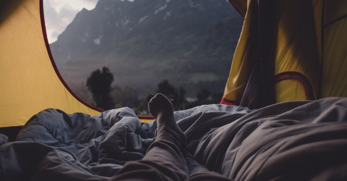 View from inside a tent with a sleeping bag and feet facing a mountain landscape at a campsite
