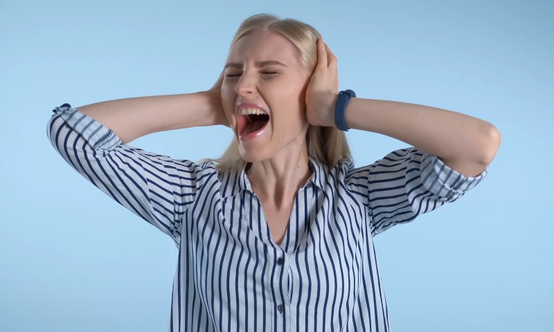 A woman in a striped shirt screams with her eyes closed, hands covering her ears