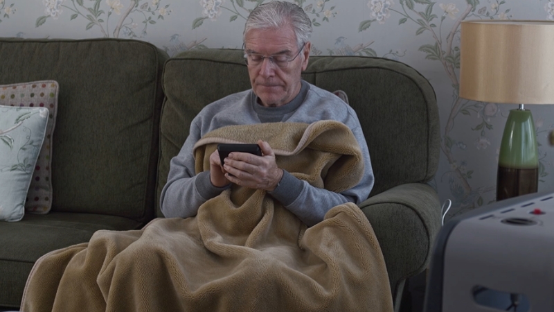 An older man sits alone on a couch, wrapped in a blanket and checking his phone