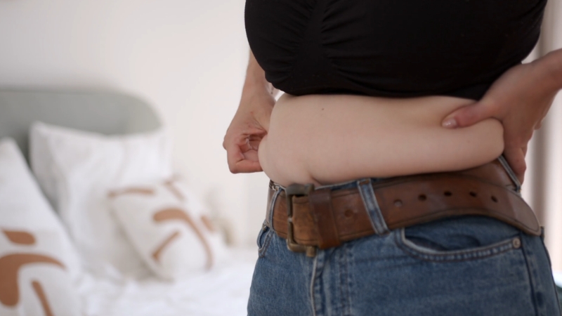 Person holding abdominal fat while standing in a bedroom, illustrating excess body weight