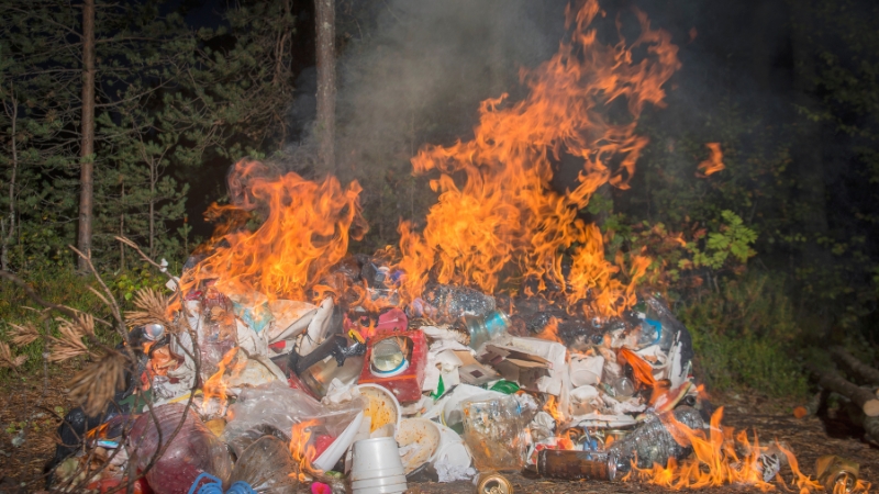 Plastic trash burns in an open fire, releasing thick smoke and visible flames in a wooded area