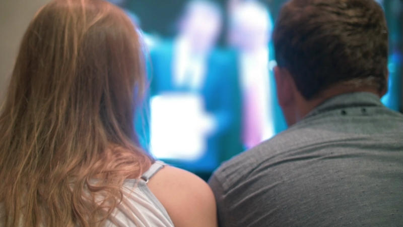 Two people sit on a couch watching traditional television in a dimly lit room
