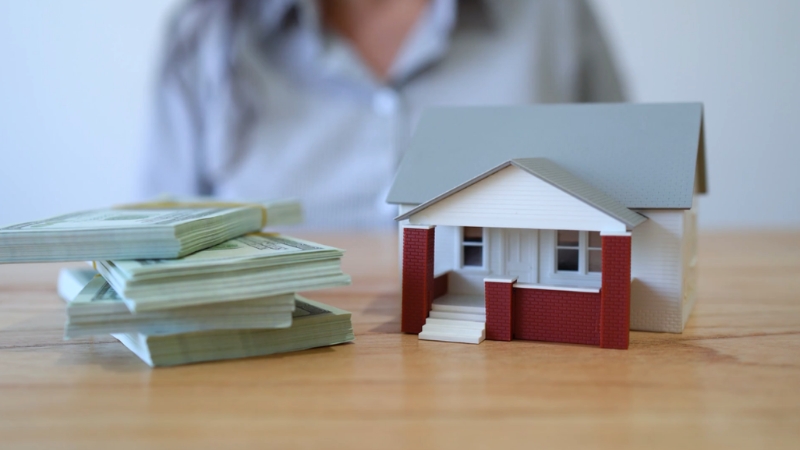 Stacks of cash sit beside a small house model, highlighting falling rents without real relief for many renters