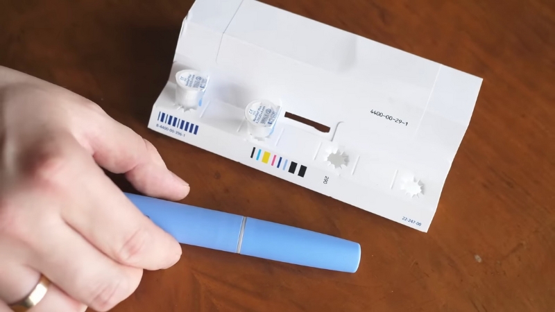 Person holding a blue injection pen next to medical test strips and packaging